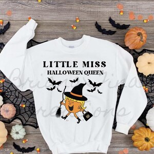 以下が含まれることがあります： 「Little Miss Halloween Queen」という文字と、ほうきとカボチャを持った魔女の漫画イラストが描かれた白い長袖スウェットシャツです。