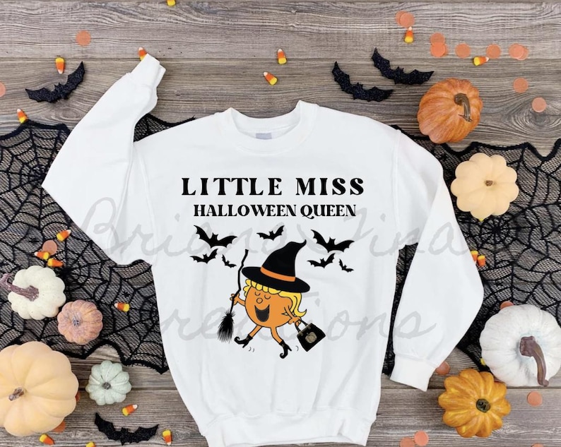 以下が含まれることがあります： 「Little Miss Halloween Queen」という文字と、ほうきとカボチャを持った魔女の漫画イラストが描かれた白い長袖スウェットシャツです。