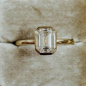 Può includere: Un anello d'oro con un diamante taglio smeraldo incastonato in una montatura a castone. L'anello è presentato in un portagioie di velluto grigio. Il diamante ha una forma rettangolare con un motivo di sfaccettature a gradini.