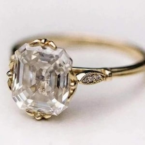May include: A gold ring with a large, clear, emerald-cut gemstone. The ring has a delicate, ornate design around the gemstone and along the band, with small accent stones near the center. The ring is set against a white background.