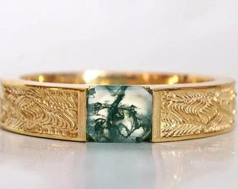 Puede incluir: Un anillo de banda de oro con una piedra preciosa de ágata verde musgo engastada en el centro. La banda está texturizada con un patrón ondulado.