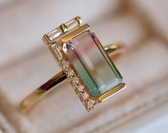 May include: A gold ring with a rectangular green, pink, and blue gemstone surrounded by a border of small diamonds. The ring is set in a gold band.