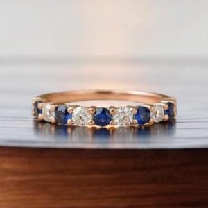 以下が含まれることがあります： ローズゴールドのバンドリング。丸いブルーサファイアとクリアダイヤモンドが交互に並んでいます。リングは、水上の夕日のぼやけた背景を持つ木製の表面にセットされています。