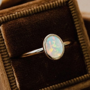Può includere: Un anello d'oro con una pietra di opale ovale incastonata in una fascia semplice. L'anello è esposto in una scatola per anelli in velluto marrone.
