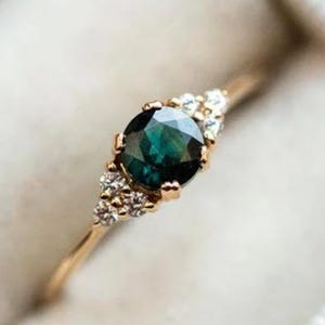 Può includere: Un anello d'oro con una grande gemma rotonda verde scuro come punto focale. Piccole gemme chiare affiancano la pietra principale su entrambi i lati. L'anello è posto in un cofanetto bianco strutturato.