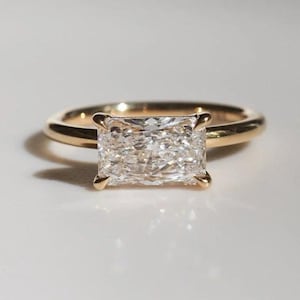 May include: A gold ring featuring a rectangular, clear gemstone. The ring has a thin, polished band, and the gemstone is secured with four prongs. The ring is presented on a white surface.