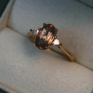 May include: A gold ring with an oval, orange-brown gemstone as the centerpiece, flanked by two smaller, white triangular stones. The ring is set in a dark gray jewelry box with a white interior.