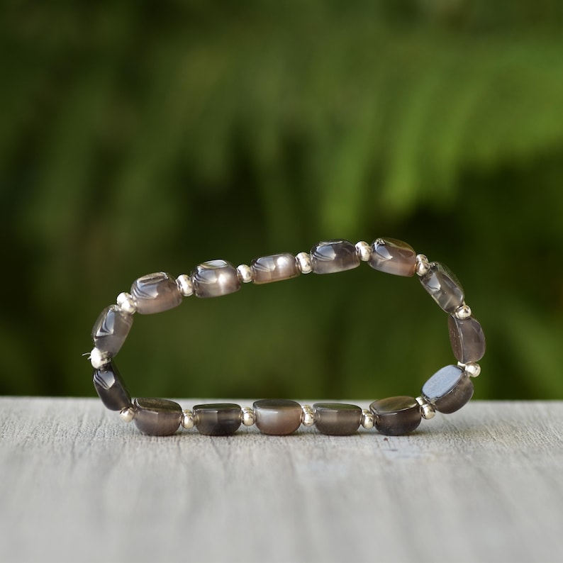 Puede incluir: Una pulsera con cuentas ovaladas de color gris-marr&oacute;n y peque&ntilde;as cuentas plateadas. La pulsera se muestra sobre una superficie de madera, con un fondo verde borroso. Las cuentas miden aproximadamente 0,8 cm de largo.