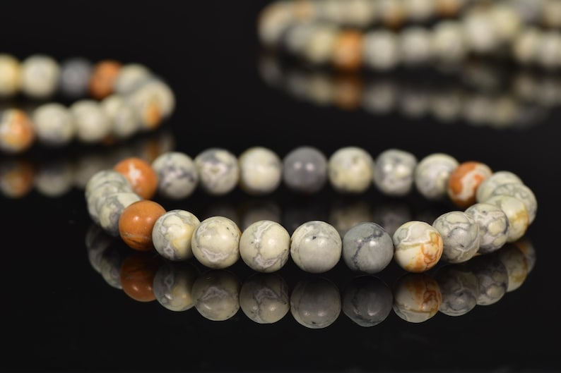 K&ouml;nnte beinhalten: Ein Armband aus runden, mehrfarbigen Steinen. Die Steine sind eine Mischung aus Wei&szlig;, Grau und Braun mit einem marmorierten Muster. Das Armband liegt auf einer schwarzen Oberfl&auml;che.