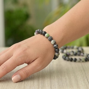 Puede incluir: Un brazalete de cuentas redondas en tonos gris oscuro, gris claro y verde. El brazalete se lleva en la muñeca. Un segundo brazalete es visible en el fondo. Las cuentas tienen un aspecto moteado.