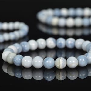 Op de afbeelding: Drie armbanden van blauwe en witte kralen worden getoond op een reflecterend zwart oppervlak. De ronde, gepolijste kralen zijn in tinten lichtblauw en wit. De armbanden zijn cirkelvormig, met de kralen aan elkaar geregen.