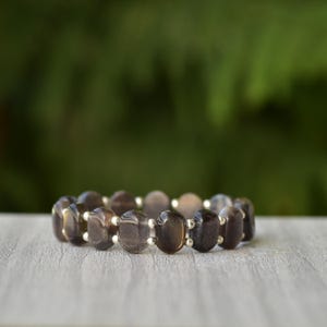 Puede incluir: Una pulsera de cuentas con piedras preciosas ovaladas de color marr&oacute;n oscuro. Las piedras est&aacute;n unidas con peque&ntilde;as cuentas plateadas. La pulsera se muestra sobre una superficie de madera clara con un fondo verde borroso.