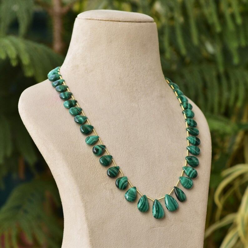 May include: A necklace featuring teardrop-shaped malachite beads strung on a gold-colored chain. The beads are a deep green with darker green veining, displayed on a beige mannequin against a blurred green background.