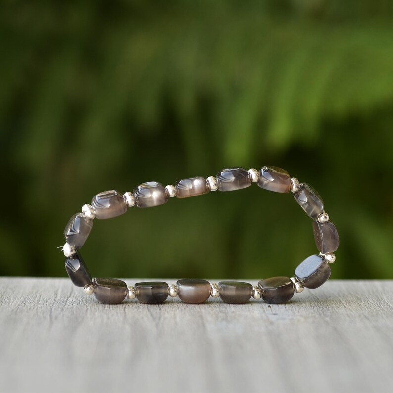 Puede incluir: Una pulsera con piedras preciosas ovaladas de color gris-marr&oacute;n ensartadas con peque&ntilde;as cuentas plateadas. La pulsera se muestra sobre una superficie de madera con un fondo verde borroso. Las piedras miden aproximadamente 1 cm de largo.