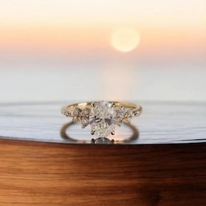 Puede incluir: Un anillo de oro con una piedra central de diamante de talla ovalada. El anillo presenta diamantes de acento más pequeños y una banda delicada. El anillo está engastado sobre un fondo borroso de una puesta de sol sobre el agua.