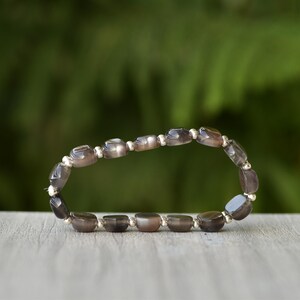 Puede incluir: Una pulsera con cuentas ovaladas de color gris-marr&oacute;n y espaciadores plateados. Se muestra sobre una superficie de madera, con un fondo verde borroso. Las cuentas tienen un acabado pulido. El dise&ntilde;o sugiere una est&eacute;tica natural y terrosa.