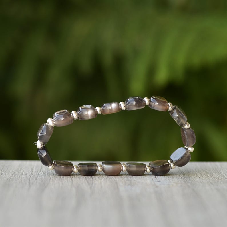 Puede incluir: Una pulsera con cuentas ovaladas de color gris-marr&oacute;n y espaciadores plateados. Se muestra sobre una superficie de madera, con un fondo verde borroso. Las cuentas tienen un acabado pulido. El dise&ntilde;o sugiere una est&eacute;tica natural y terrosa.