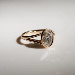 Könnte beinhalten: Ein goldener Ring mit einem großen, oval geformten Diamanten. Der Ring hat ein geteiltes Schaftdesign, wobei sich das Band in zwei separate Stränge aufteilt, wenn es sich dem Diamanten nähert. Der Diamant ist in einer Lünettenfassung gefasst.