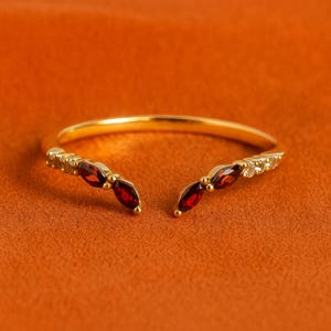 Puede incluir: Un anillo abierto de color dorado con una fila de pequeñas piedras transparentes y tres gemas rojas oscuras en forma de marquesa a cada lado. El anillo está sobre un fondo naranja.