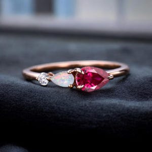 May include: Rose gold ring with a cluster of gemstones. It features a round, clear stone, a pear-shaped opal, and a vibrant, pear-shaped ruby. The ring is set against a dark background.