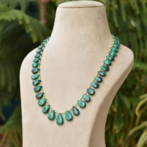 May include: A green malachite necklace displayed on a mannequin. The necklace features teardrop-shaped beads strung on a gold-colored chain. The beads have a marbled appearance, with varying shades of green. The necklace is a jewelry item.