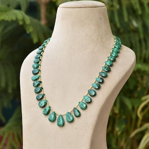 May include: A necklace featuring polished, teardrop-shaped malachite beads in varying shades of green. The beads are strung on a delicate gold-colored chain, displayed on a neutral-toned mannequin. The necklace is a piece of jewelry.