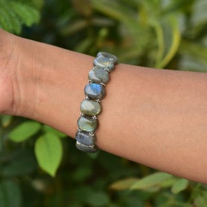 Puede incluir: Un brazalete de labradorita con piedras ovaladas en tonos grises, verdes y azules. Las piedras están unidas con pequeñas cuentas plateadas. El brazalete se lleva en la muñeca.
