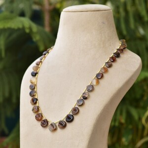 Puede incluir: Un collar con cuentas pulidas, planas y redondas en tonos marr&oacute;n, beige y gris. Las cuentas est&aacute;n ensartadas en un fino alambre dorado. El collar se exhibe sobre un busto de maniqu&iacute; beige sobre un fondo verde borroso.