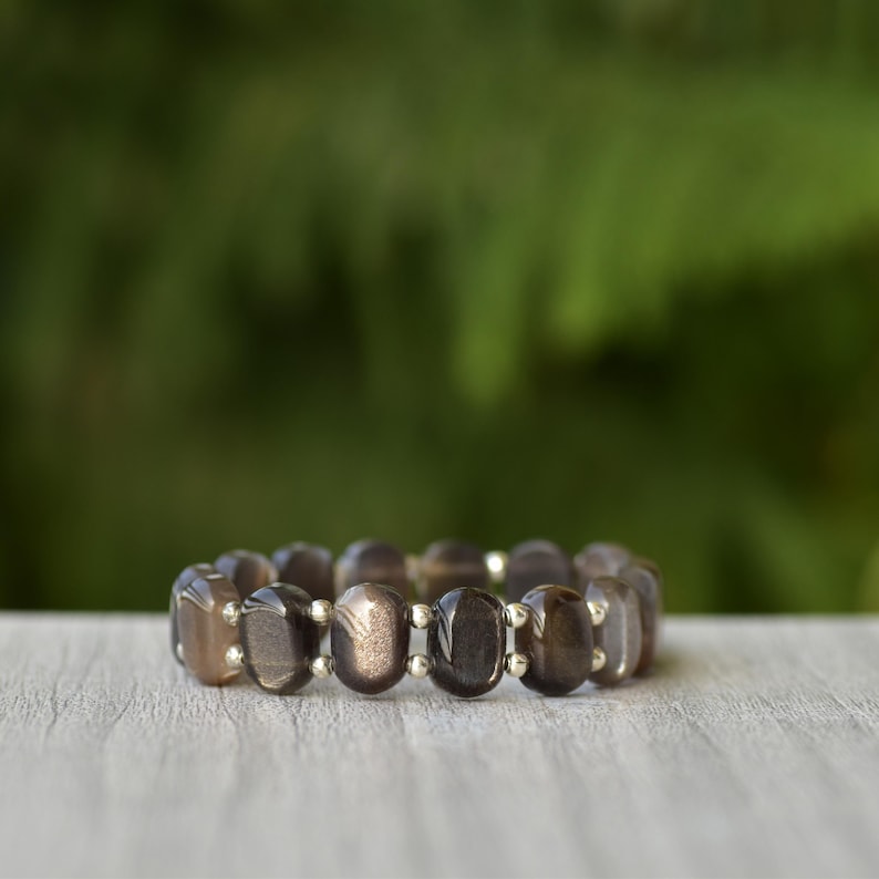Puede incluir: Una pulsera con cuentas ovaladas de color marr&oacute;n oscuro con un brillo sutil. Las cuentas est&aacute;n conectadas por peque&ntilde;os eslabones met&aacute;licos plateados. La pulsera se exhibe sobre una superficie de madera clara con un fondo verde borroso.