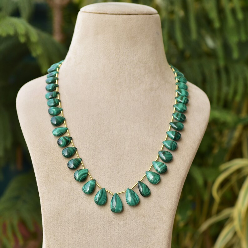 May include: A necklace featuring teardrop-shaped malachite beads in varying shades of green. The beads are strung on a delicate gold-colored chain, displayed on a neutral-toned mannequin. The background is a soft blur of green foliage.