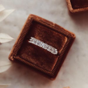 Puede incluir: Un anillo de plata con múltiples diamantes pequeños de corte rectangular colocados en fila. El anillo se muestra dentro de un joyero cuadrado de terciopelo marrón. El fondo es de un color suave y neutro.