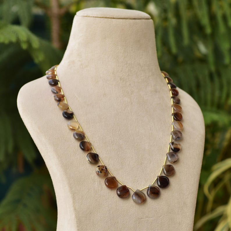 Puede incluir: Un collar con cuentas pulidas en forma de l&aacute;grima en tonos marr&oacute;n, beige y gris. Las cuentas est&aacute;n ensartadas en una delicada cadena dorada, expuestas sobre un maniqu&iacute; beige. El fondo es verde borroso.