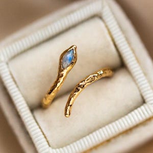 Puede incluir: Anillo de serpiente de oro con una piedra de luna. El anillo es texturizado y tiene un diseño delicado.