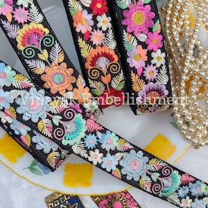 May include: Four black ribbon trims with colorful floral embroidery. The trims are on a white plate with a pearl necklace and two decorative tassels.