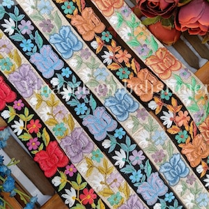 May include: Five different embroidered trims with floral designs. The trims are in various colors, including black, white, orange, blue, and red. The trims are arranged in a row on a wooden surface. emb1360