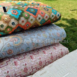 May include: Four different fabric swatches stacked on top of each other. The top swatch is a colorful floral pattern with sequins and mirror embellishments. The second swatch is a light blue fabric with a floral pattern. The third swatch is a pink and white fabric with a geometric pattern. The bottom swatch is a white fabric with a geometric pattern. The text "Embf061" is visible on the bottom swatch.