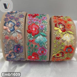 Puede incluir: Tres rollos de ribete bordado con diseños florales. El ribete presenta una base de malla beige con flores coloridas en tonos de azul, rojo, naranja y morado. El texto "Emb1609" es visible en la parte inferior.