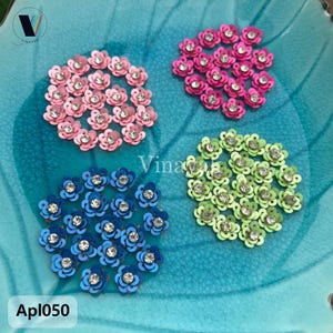 Puede incluir: Cuatro grupos de adornos florales con lentejuelas en rosa, fucsia, azul y verde, cada uno con un pedrería central. Las flores están dispuestas sobre una superficie turquesa. El texto "Apl050" es visible.