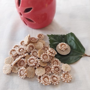 Puede incluir: Un montón de adornos redondos de tela beige con ribete dorado y cuentas blancas. Los adornos están esparcidos sobre una superficie blanca.