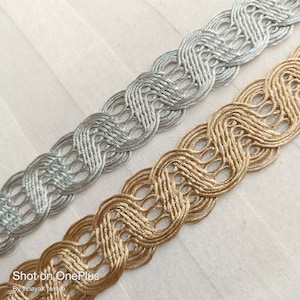 May include: Two braided trim cords, one silver and one gold, with a decorative looped pattern.  The cords are lying on a white fabric surface.  Shot on OnePlus. By vinayak textile.