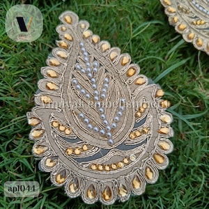 May include: A gold and white embroidered fabric applique in the shape of a teardrop with a paisley design. The applique is decorated with gold sequins and white pearls.