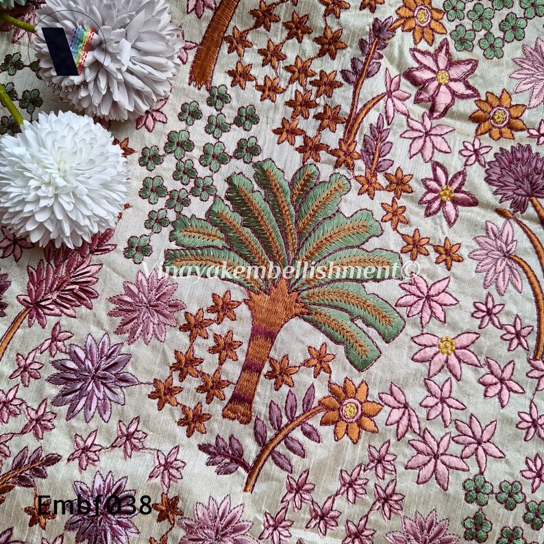May include: A close-up of a cream-coloured fabric with intricate embroidery of pink, green, and orange flowers and trees. The fabric has a subtle texture and the embroidery is detailed and delicate.