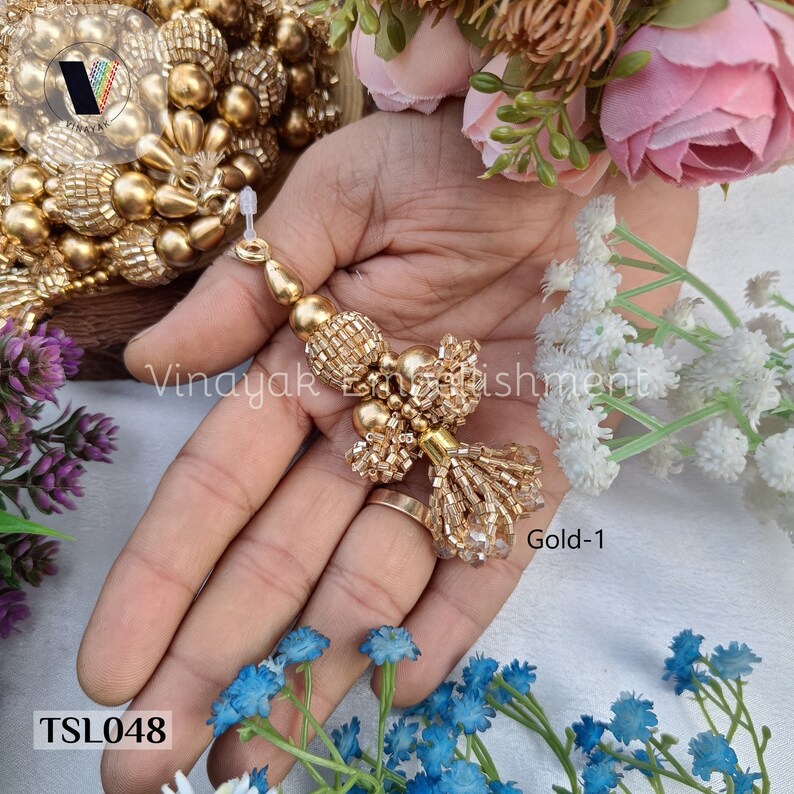 May include: Gold beaded tassel trim with a gold and clear beaded fringe. The trim is shown on a hand with a ring. The trim is labeled "Gold-1" and "TSL048".