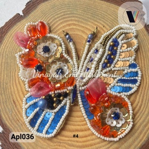 May include: A colorful butterfly-shaped beaded applique with gold, blue, white, and orange sequins. The applique is on a wooden background and has the text "Apl036 #4" below it.
