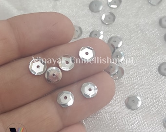 May include: Silver sequins with a hexagonal shape. The sequins are scattered on a white fabric surface.