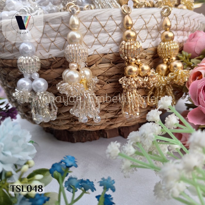 May include: Three sets of decorative tassels in a wicker basket. The tassels are made of beads and have a pearl-like finish. The tassels are white, gold, and silver.
