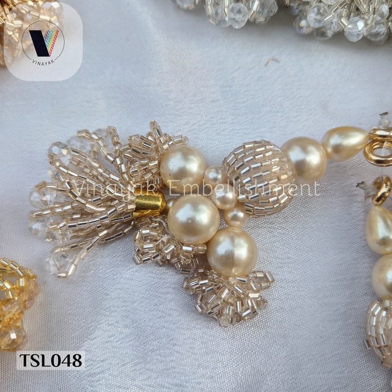 May include: A close-up of a white beaded tassel with a gold accent. The tassel is made of small, round beads and has a pearl-like finish. The tassel is attached to a string of pearls and beads. The text "TSL048" is visible in the lower left corner of the image.