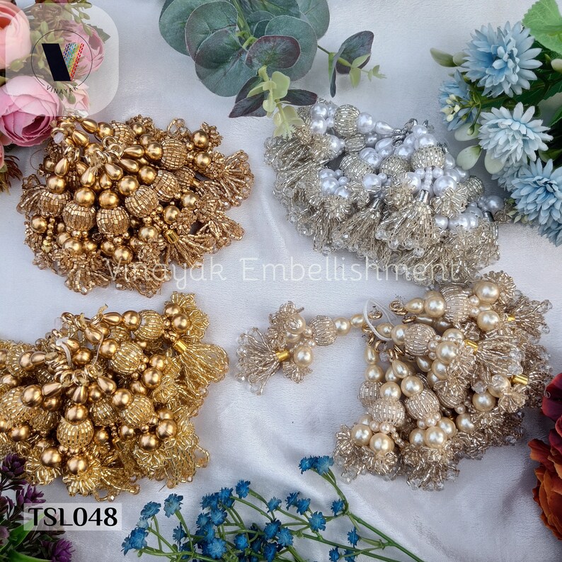 May include: Four sets of beaded tassels in gold, white, and cream colors. The tassels are made of small beads and have a fringe-like appearance. The tassels are arranged on a white surface with flowers in the background. The text "TSL048" is visible in the bottom left corner of the image.