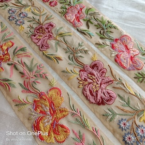 Puede incluir: Tres galones de tela beige con diseños florales bordados en tonos de rosa, rojo, amarillo, verde y azul. El galón presenta un patrón repetitivo de flores y hojas.