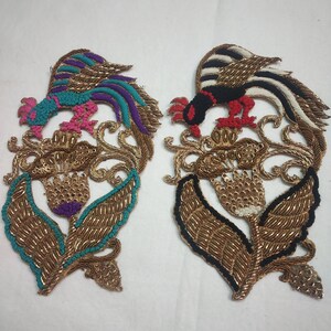 May include: Two embroidered patches featuring a stylized bird perched on a floral design. The bird on the left is blue, purple, and green, while the bird on the right is black, white, and red. Both patches have gold thread embroidery and sequins.